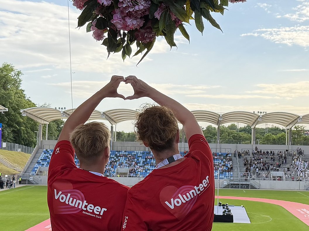 Zwei Personen in roten T-Shirts mit der Aufschrift 'Volunteer' formen mit den Händen ein Herz vor einer Stadiontribüne